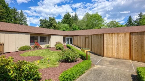 A central courtyard with a grassy area, a wooden walkway, a concrete path, and a building with a mix of tan siding and natural wood paneling
