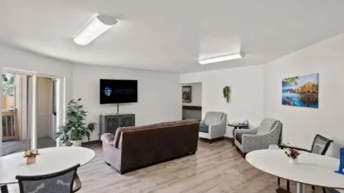A communal living area with a television, couches, and round dining tables on a light wood floor, with a sliding glass door leading to a deck