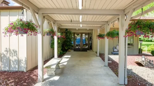 A covered walkway leading to the entrance of a building, with hanging flower baskets and a concrete bench
