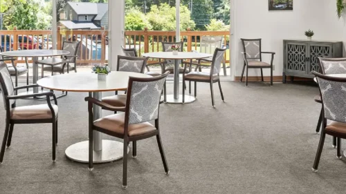 A cropped view of a dining room with round tables, chairs with patterned backs, and large windows looking out onto a deck