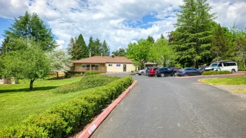 A parking lot with a paved driveway, surrounded by a grassy lawn, a hedge, and a tan-sided building with a deck