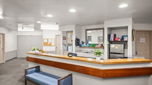 A reception desk with a light wood counter, a blue bench, a mini-refrigerator, and a mirror on the back wall
