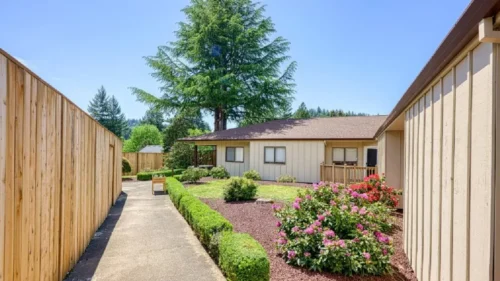 A walkway between a wooden fence and a tan-sided building, with a garden area and a bench
