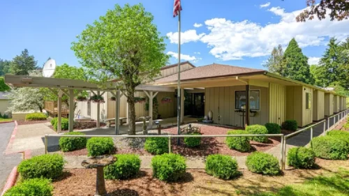 The front of a tan building with a covered entrance, an American flag on a pole, and a birdbath surrounded by bushes